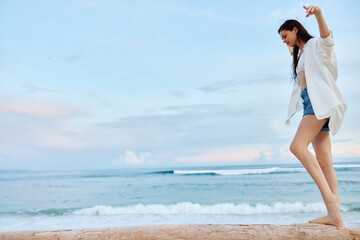 Happy tanned woman in white swimsuit shirt and denim shorts walks the beach on a log on the sand by the ocean with wet hair after swimming, sunset light in Bali