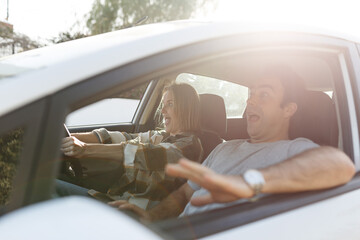 Naklejka premium Emotional millennial couple having fun while car trip, listening to music and cheerfully singing, shot from inside the auto, sun flare.
