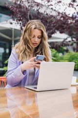 Sexy blonde in a stylish suit types a message on her phone while working outside. Breakfast on the summer terrace