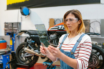 Concentrated female mechanic with security glasses using transparent digital tablet with diagnosis app to review motorcycle on factory
