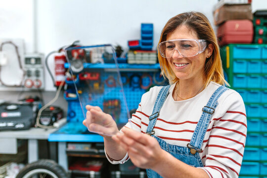 Portrait of happy female mechanic with security glasses touching transparent tablet while working on garage