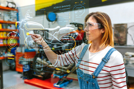 Portrait of young female mechanic with glasses using hud panel screen with augmented reality hologram to review motorcycle on factory. Futuristic user interface concept.