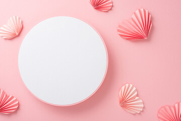 Top view photo of empty white circle with a pastel pink ribbon and paper origami hearts on an isolated light pink background with copyspace