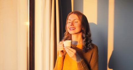 Portrait of happy satisfied smiling young woman enjoying hot tasty cup of coffee or tea in early morning at home by window with sunlight