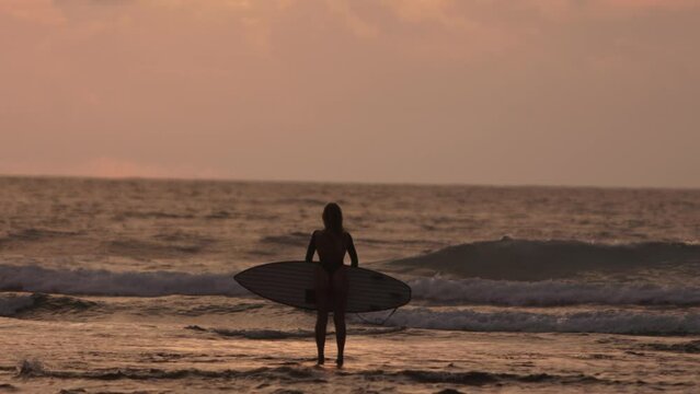 Rear back view silhouette of person surfer stands in sunset evening sea allowing waves to wash his feet, giving feeling of freedom, lightness. Concept of freedom easy attitude to life without worries.