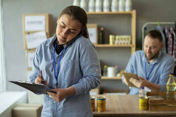 Young woman wearing volunteer uniform talking on the smartphone writing on clipboard at charity center. Volunteers working in distribution or refugee assistance center