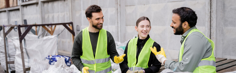Cheerful multiethnic sorters in high visibility vests and gloves talking while standing near trash in outdoor waste disposal station, garbage sorting and recycling concept, banner