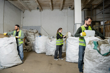 Multiethnic workers in high visibility vests and gloves separating trash in sacks while working in...