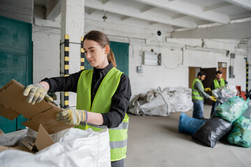 Young female worker in protective vest and gloves holding cardboard near sack and blurred...