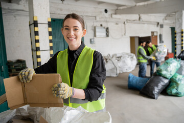 Cheerful young worker in protective vest and gloves holding cardboard and looking at camera while...