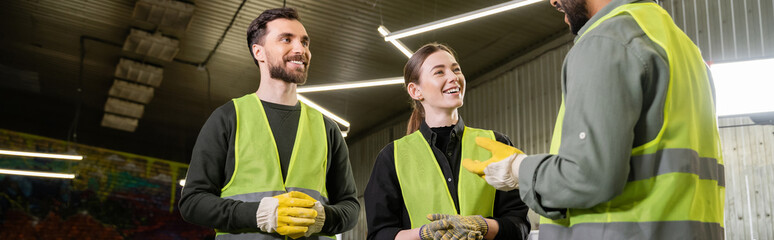 Cheerful workers in protective gloves and high visibility vests looking at indian colleague talking...
