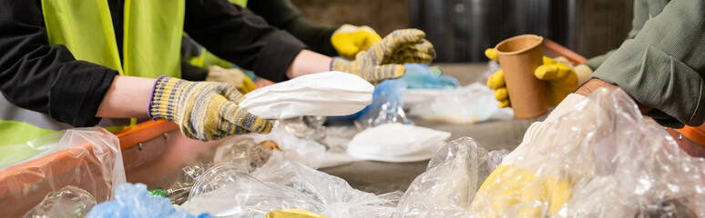 Cropped view of workers in protective gloves taking different plastic and paper trash from conveyor while working together in waste disposal station, recycling concept, banner