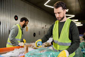 worker in protective gloves and vest putting tin can in plastic bag while separating trash near...