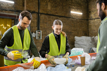 Positive young worker in gloves and protective clothes sorting garbage on conveyor near multiethnic...