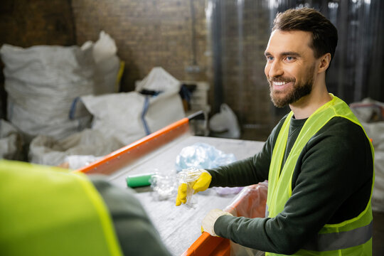 Cheerful male sorter in protective vest and gloves looking at blurred colleague while sorting garbage near conveyor in waste disposal station, garbage sorting and recycling concept
