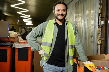 Fototapeta Smiling indian worker in high visibility vest and protective gloves looking at camera near plastic caps in carton boxes while working in waste disposal station, garbage sorting and recycling concept obraz