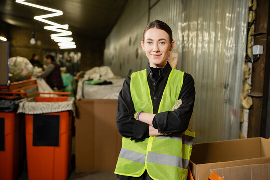 Young and smiling worker of waste disposal station in high visibility vest looking at camera and crossing arms while standing near blurred bins at background, garbage recycling concept