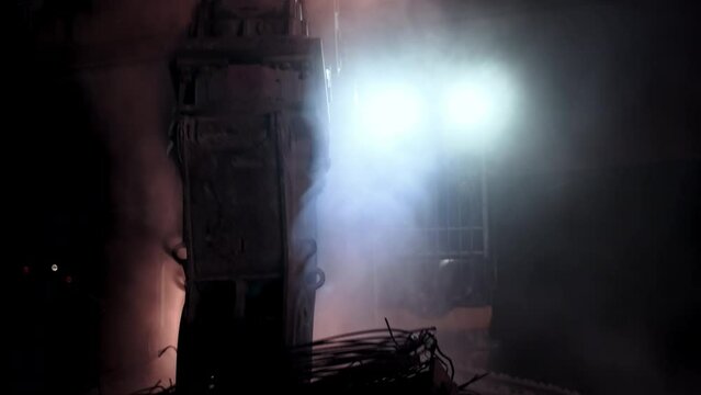Bunch of steel rebars picked up from ground with hydraulic claw on excavator, night time with lights illuminating demolition site