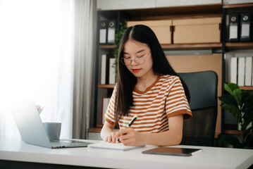Fototapeta na wymiar Young attractive Asian woman smiling thinking planning writing in notebook, tablet and laptop working from home, at home office