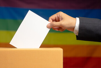 Hand voter holding ballot paper putting into the voting box at place election against a rainbow flag background