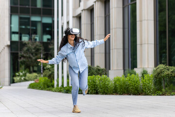 Young beautiful girl in shirt and jeans dancing outside office building wearing virtual reality glasses, hispanic woman happy having fun and relaxing with curly hair.