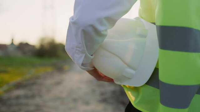 Close View of Engineer Man Hands Holding Construction Helmet on the Background of the Construction Site. Developer or Contractor Inspects the Construction Progress. Protective Hard Hat