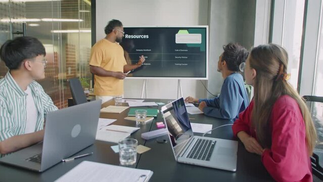 Young businessman giving presentation on interactive digital screen to ethnically diverse group of colleagues during office meeting