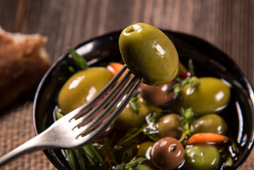 Pricked olive on a fork against the background of olives in a black bowl, on a wooden table.