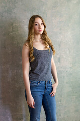Beautiful slim young woman with long wavy blond hair standing relaxed in tank top and jeans against concrete wall