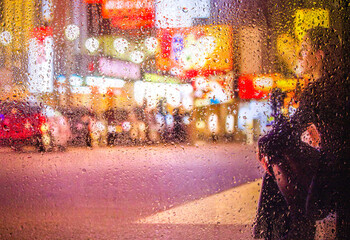 View through a glass window with raindrops on a blurred silhouette of a people walking on autumn rain , night street scene. focus on raindrops	