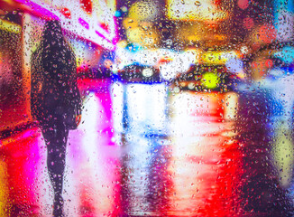 View through glass window with rain drops on blurred reflection silhouettesof a girl in walking on a rain under umbrellas and bokeh city lights, night street scene. Focus on raindrops on glass	