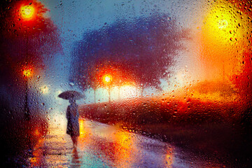View through a glass window with raindrops on the silhouette of a man walking under an umbrella down the street in the morning mist in the rain Colorful lights of street lamps Focus on the raindrops
