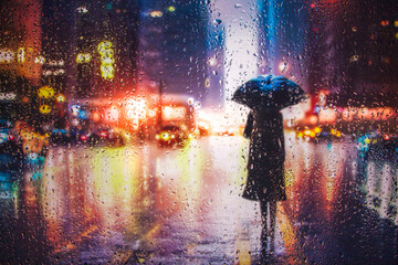 View through glass window with rain drops on blurred reflection silhouette of a girl on a city street after rain and colorful neon bokeh city lights, night street scene. Focus on raindrops on glass 