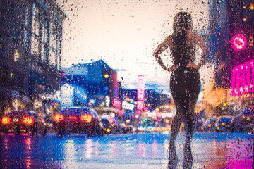View through glass window with rain drops on blurred reflection silhouette of a girl on a city street after rain and colorful neon bokeh city lights, night street scene. Focus on raindrops on glass