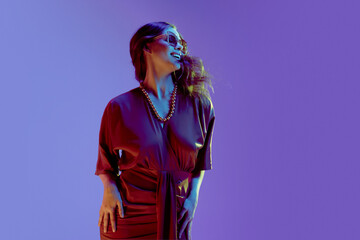Happy fashion model, girl wearing elegant silk red dress with necklace and sunglasses smiling over gradient purple background in neon light. Latest trends