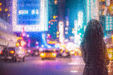View through glass window with rain drops on blurred reflection silhouette of a girl on a city...