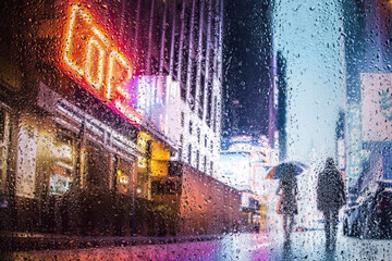 View through glass window with rain drops on blurred  silhouette of a girl  under an um on a city street after rain and colorful neon bokeh city lights, night street scene. Focus on raindrops on glass