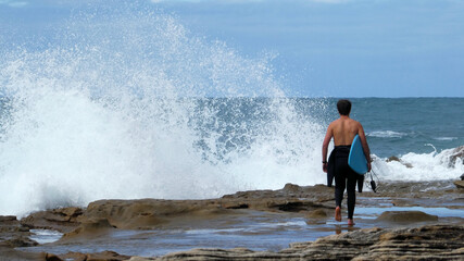 rear view of young man carrying surfboard walking towards huge wave splashing onto rocks