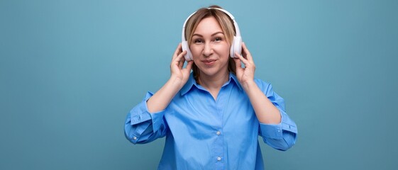 horizontal photo of a smiling young woman in a casual shirt listening to music in big white headphones on a blue isolated background with copy space