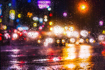 View through a glass window with raindrops on city streets with cars in the rain, bokeh of colorful city lights, night street scene. Focus on raindrops on glass