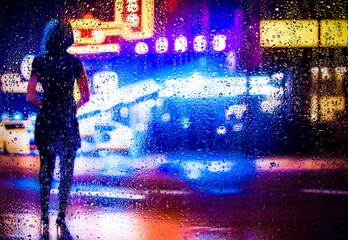 View through glass window with rain drops on blurred reflection silhouette of a girl on a city street after rain and colorful neon bokeh city lights, night street scene. Focus on raindrops on glass