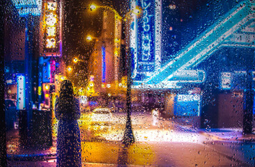 view through glass window with rain drops on blurred reflection silhouette of a girl on a city street after rain and colorful neon bokeh city lights, night street scene. Focus on raindrops on glass	