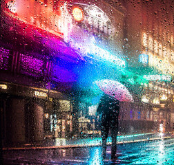 View through glass window with rain drops on blurred reflection silhouette of a man on a city street after rain and colorful neon bokeh city lights, night street scene. Focus on raindrops on glass