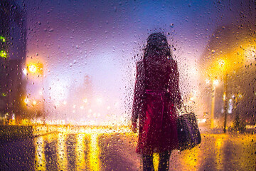 View through glass window with rain drops on blurred reflection silhouette of a girl on a city street after rain and colorful neon bokeh city lights, night street scene. Focus on raindrops on glass