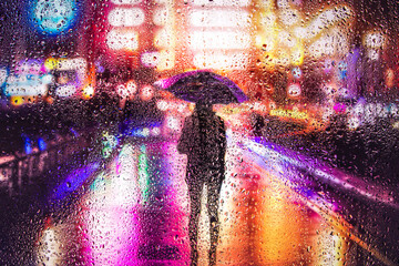 View through glass window with rain drops on blurred reflection silhouette of a man on a city street after rain and colorful neon bokeh city lights, night street scene. Focus on raindrops on glass