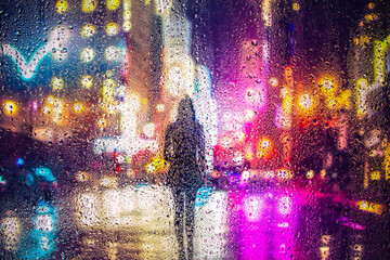 View through glass window with rain drops on blurred reflection silhouette of a girl on a city street after rain and colorful neon bokeh city lights, night street scene. Focus on raindrops on glass