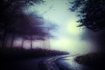 View through a glass window with raindrops on the silhouette of a man walking under an umbrella down the street in the morning mist in the rain. Colorful lights of street lamps. Focus on the raindrops