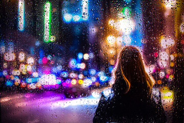 View through a glass window with raindrops on a blurred silhouette of a girl on a city street after...