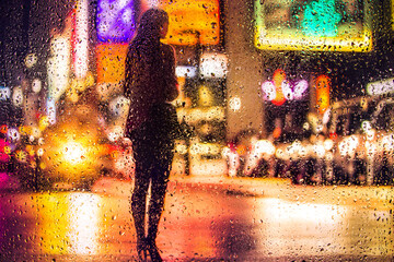 View through glass window with rain drops on blurred reflection silhouettesof a girl in walking on a rain under umbrellas and bokeh city lights, night street scene. Focus on raindrops on glass