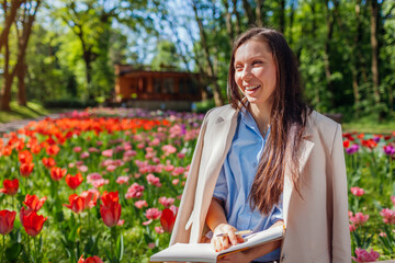 Young caucasian businesswoman laughing holding notebook in spring park. Teacher having break outdoors. Space